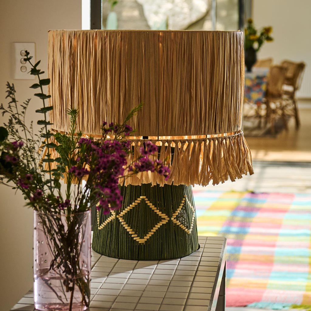 The image shows a stylish and modern lamp with a unique design. The lamp features a base made of raffia, giving it a natural, textured appearance. The raffia is woven in a way that creates an intricate pattern, adding to the lamp's aesthetic appeal. The lampshade is likely made of a light fabric, complementing the earthy tones of the raffia base. The overall look of the lamp is both contemporary and bohemian, making it a versatile piece for various interior design styles.