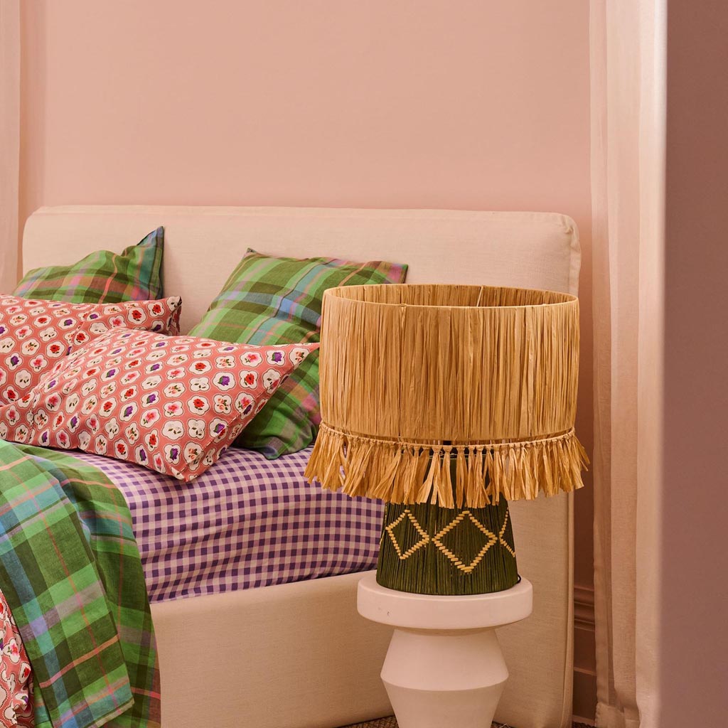 The image shows a stylish table lamp with a unique design. The lamp features a base made of natural raffia material, giving it a textured and earthy appearance. The lampshade is likely made of a similar material or a complementary fabric, contributing to the overall bohemian and coastal aesthetic. The lamp is set against a plain background, highlighting its intricate details and craftsmanship.