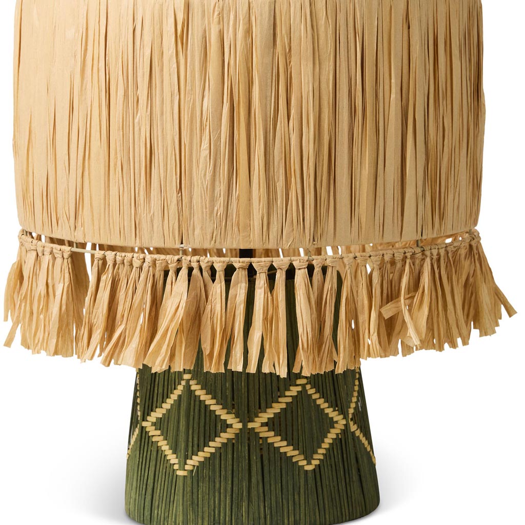 The image shows a stylish table lamp with a unique design. The lamp features a base made of natural raffia material, giving it a textured and earthy appearance. The lampshade is likely made of a light fabric, complementing the natural look of the base. The overall design of the lamp is modern yet rustic, making it a versatile piece that could fit into various interior decor styles. The lighting in the image highlights the intricate details of the raffia material, emphasizing its craftsmanship.