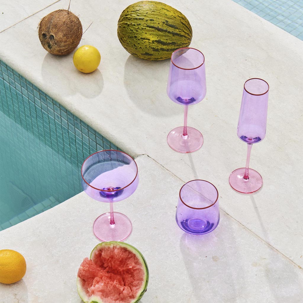 The image shows a pair of wine glasses with a unique color block design. Each glass features a lilac-colored stem and base, while the bowl of the glass is clear. The glasses are elegantly shaped, with a wide bowl that tapers slightly towards the rim. They are set against a plain, neutral background, highlighting their modern and stylish appearance.