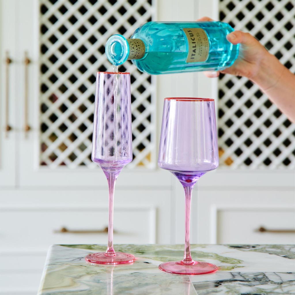 The image shows a pair of elegant wine glasses with a unique color block design. The glasses have a lilac hue at the base, transitioning into a clear glass towards the top. They are set against a plain white background, highlighting their modern and artistic design. The glasses have a sleek, slender stem and a wide bowl, typical of wine glasses, making them both functional and visually appealing.