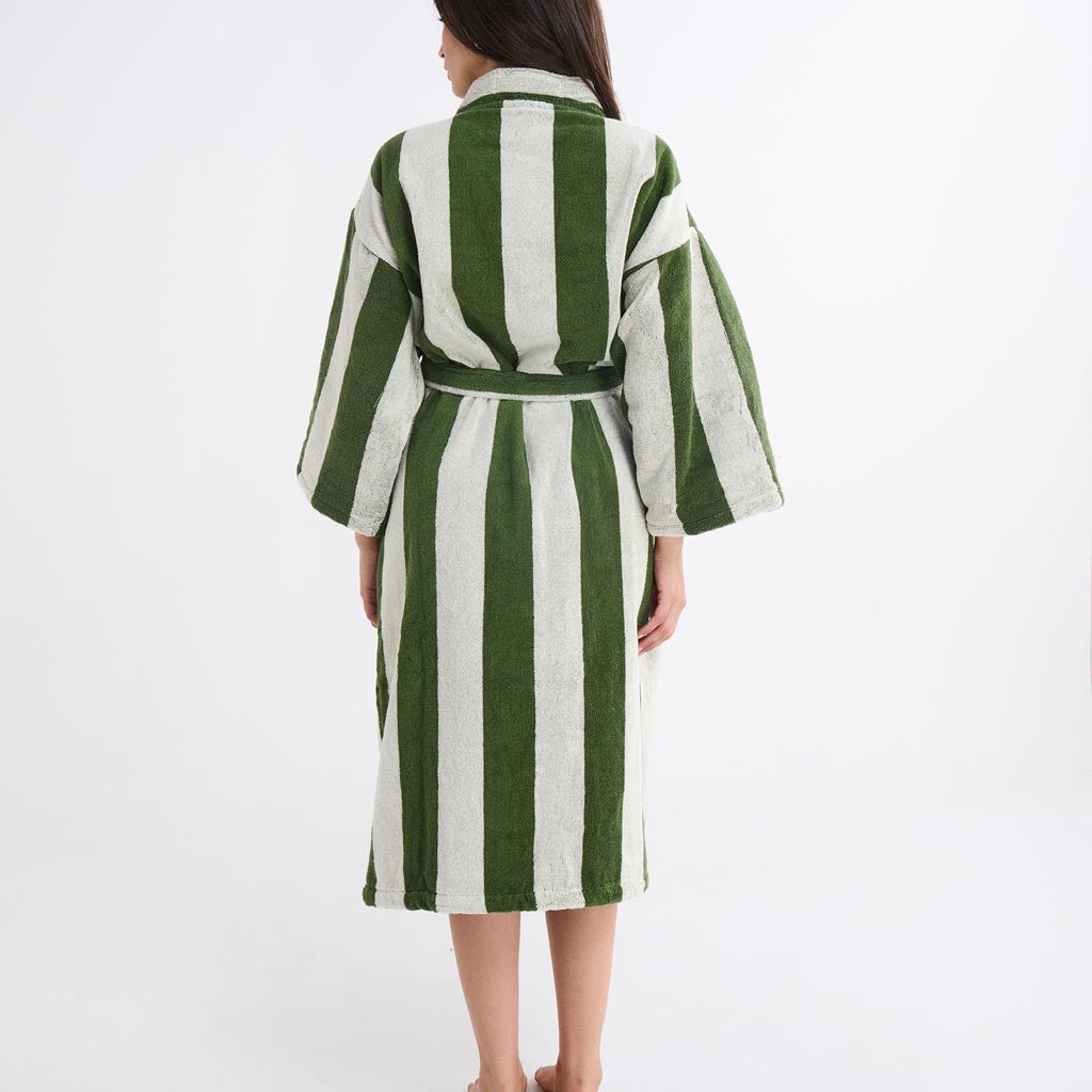 The image is of a bathrobe made by Kip & Co, featuring a design called "Moss Stripe Terry." The robe is displayed hanging against a plain background, showcasing its full length. It has a pattern of horizontal stripes in various shades of green, resembling moss, with some contrasting colors to add depth. The robe appears to be made of a soft, absorbent terry cloth material, ideal for use after bathing. It includes a belt at the waist for closure and has long sleeves, providing a cozy and comfortable fit.