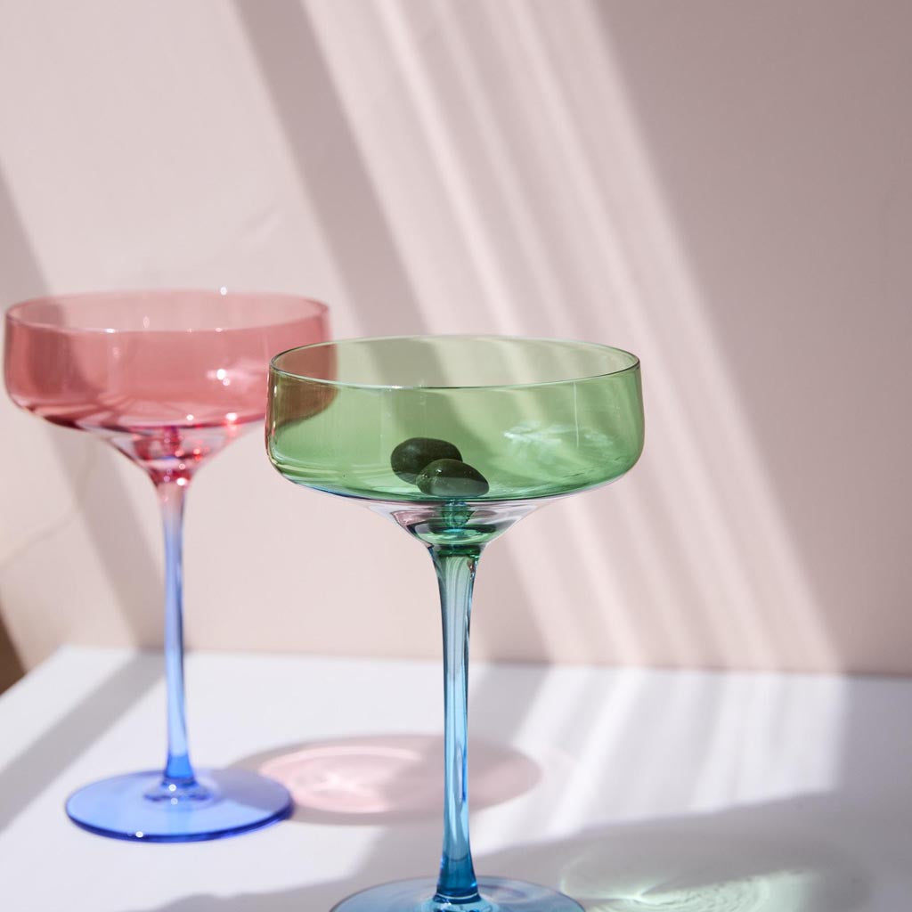 The image shows a set of two margarita glasses from Kip & Co, named "Pistachio Crush." The glasses are elegantly designed with a unique, textured surface that resembles crushed pistachios, giving them a distinctive and stylish appearance. The glassware is likely made of high-quality material, showcasing a blend of artistry and functionality, perfect for serving margaritas or other cocktails. The background is plain, ensuring the focus remains on the intricate details and vibrant color of the glasses.
