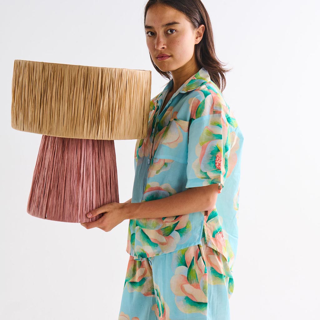 The image is of a decorative lamp made from raffia material. The lamp has a natural, earthy appearance with a woven texture, showcasing the intricate craftsmanship of the raffia fibers. The lampshade is likely in a neutral or warm tone, complementing the organic feel of the design. The lamp's base may be simple to highlight the unique texture and style of the raffia shade. This type of lamp would add a rustic or bohemian touch to interior decor.