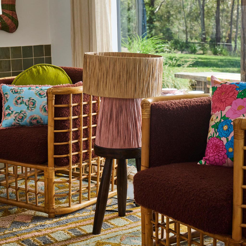 The image is of a decorative lamp made from raffia material. The lamp has a natural, earthy appearance with a woven texture, showcasing a bohemian or rustic style. The lampshade is likely crafted from strands of raffia, giving it a fringed or layered look. The color is a warm, neutral tone, possibly resembling shades of beige or light brown, which complements a variety of interior decor styles. The lamp is likely designed to add a touch of organic elegance to a room.