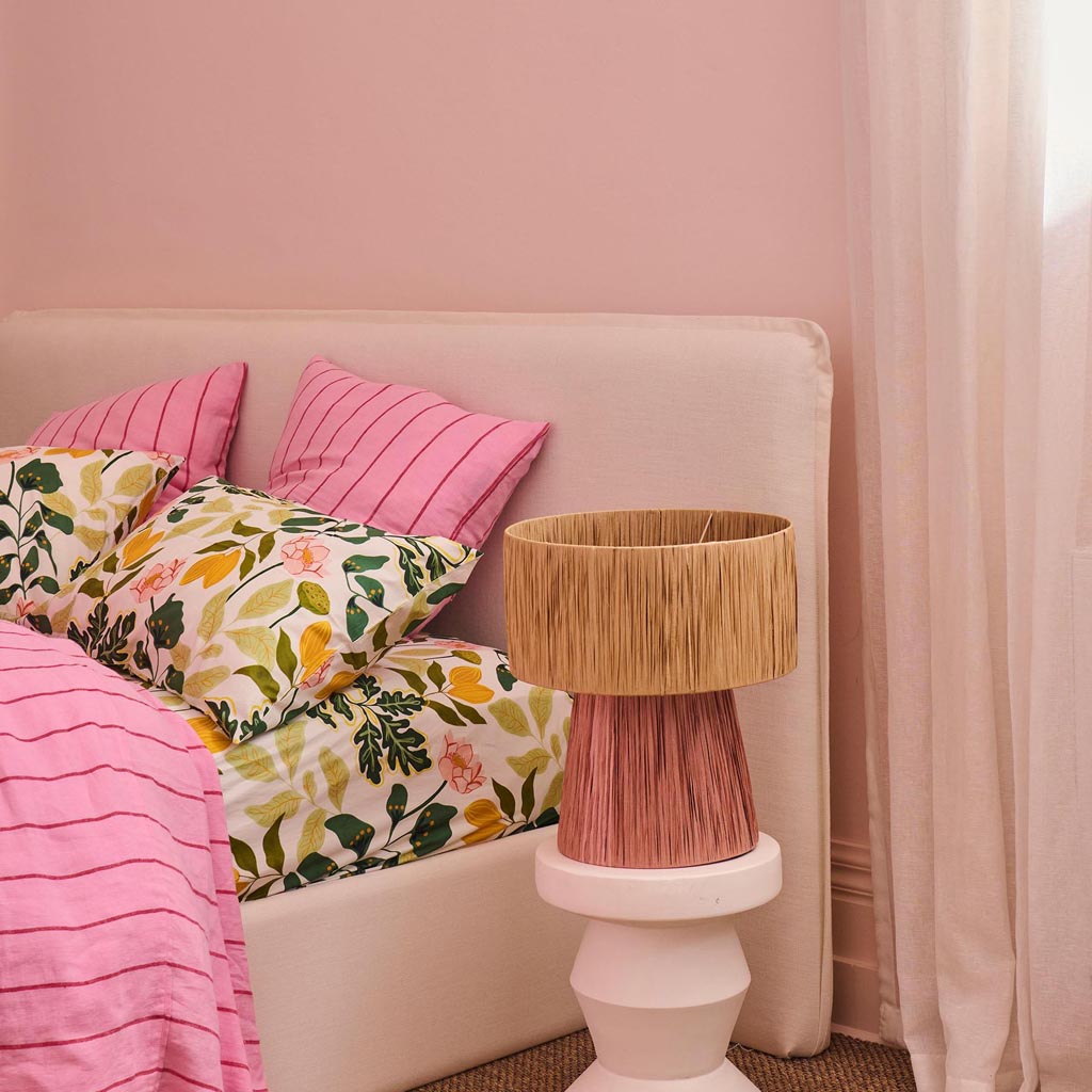 The image is of a decorative lamp made from raffia material. The lamp has a natural, earthy appearance with a woven texture, showcasing a design that is both rustic and stylish. The lampshade is likely crafted from strands of raffia, giving it a unique, handcrafted look. The color of the lamp is a warm, neutral tone, reminiscent of dried grass or straw, which complements a variety of interior design styles. The lamp is likely designed to add a touch of bohemian or natural charm to a room.