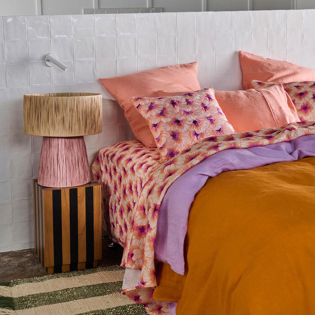 The image is of a decorative lamp made from raffia material. The lamp has a natural, earthy appearance with a woven texture, showcasing the intricate craftsmanship of the raffia fibers. The lampshade is likely in a neutral or warm tone, complementing the organic feel of the design. The lamp may be placed on a table or a flat surface, and it exudes a bohemian or rustic aesthetic, suitable for adding a touch of nature-inspired decor to a room.