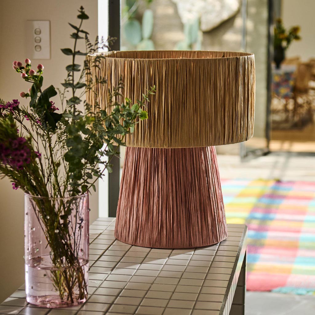 The image shows a stylish and modern lamp with a base made of raffia, a natural fiber known for its textured and woven appearance. The lamp has a cylindrical shape with a slightly tapered top, and the raffia material gives it a rustic and earthy look. The lampshade is likely made of a neutral-colored fabric, complementing the natural tones of the raffia base. The overall design is contemporary yet organic, making it suitable for a variety of interior styles, from bohemian to minimalist.