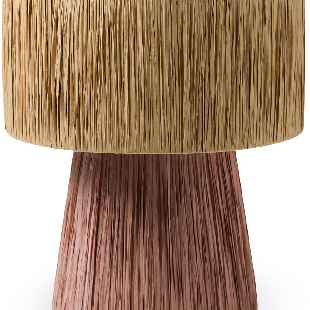 The image shows a stylish table lamp with a unique design. The lamp features a base made of natural raffia material, giving it a textured and earthy appearance. The lampshade is likely made of a similar material or a complementary fabric, contributing to the overall rustic and bohemian aesthetic. The lamp is set against a plain background, highlighting its intricate details and craftsmanship. The color palette includes warm, neutral tones, making it a versatile piece for various interior decor styles.
