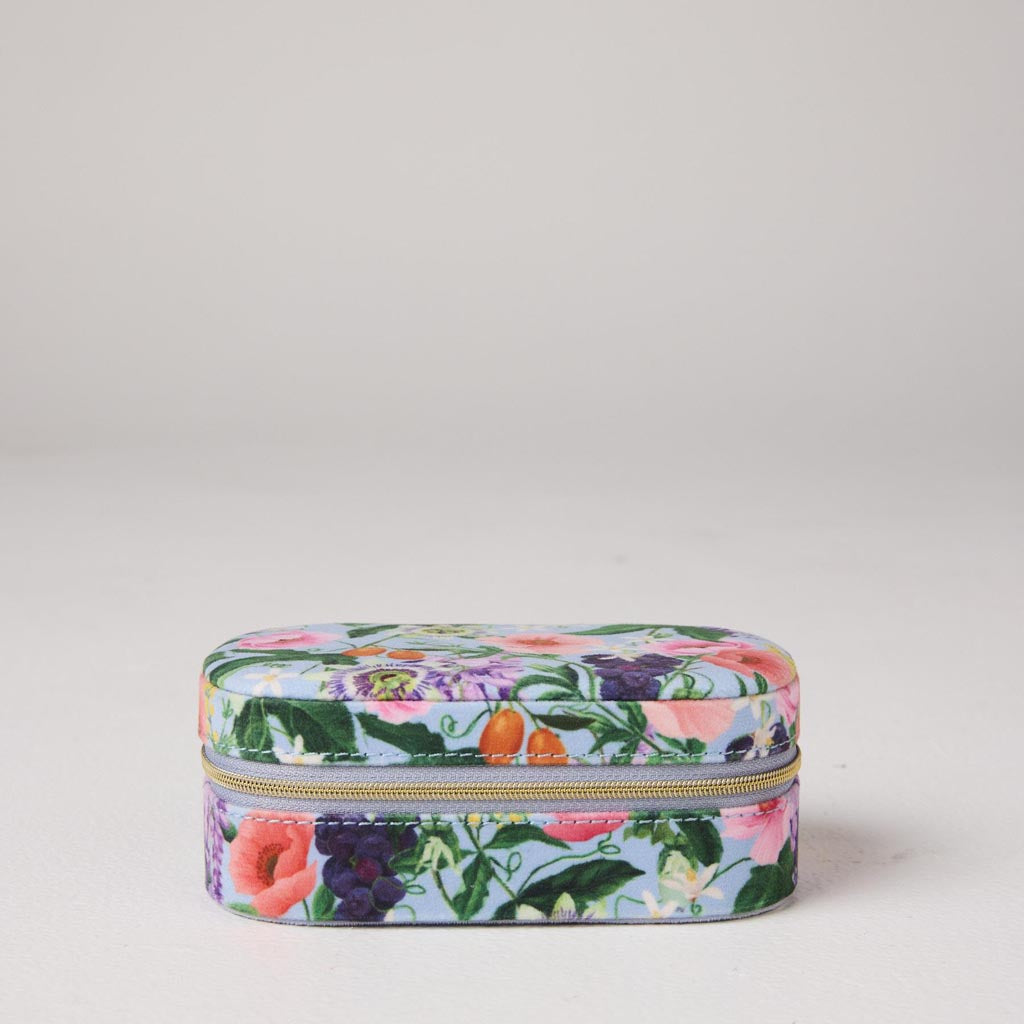 The image shows a small, rectangular velvet jewelry box in a soft pastel pink color. The box has a smooth, plush texture typical of velvet material. It features a gold-colored zipper that runs along the edge, providing a secure closure. The zipper pull is also gold and has a small, elegant design. The box is likely designed for travel, as it is compact and portable, making it convenient for storing and protecting jewelry items while on the go. The background is a simple, neutral color, allowing the jewelry 