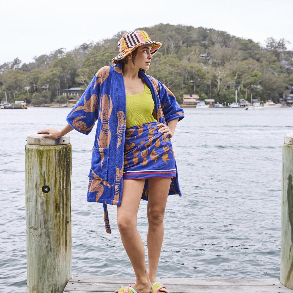 The image is of a colorful sarong made from organic cotton. The design features a vibrant pattern with a mix of warm and cool colors, including shades of orange, pink, yellow, and blue. The pattern may include abstract shapes or floral motifs, giving it a lively and summery feel. The fabric appears lightweight and soft, suitable for use as a beach cover-up or a stylish accessory. The sarong is likely displayed against a plain background to highlight its design and colors.