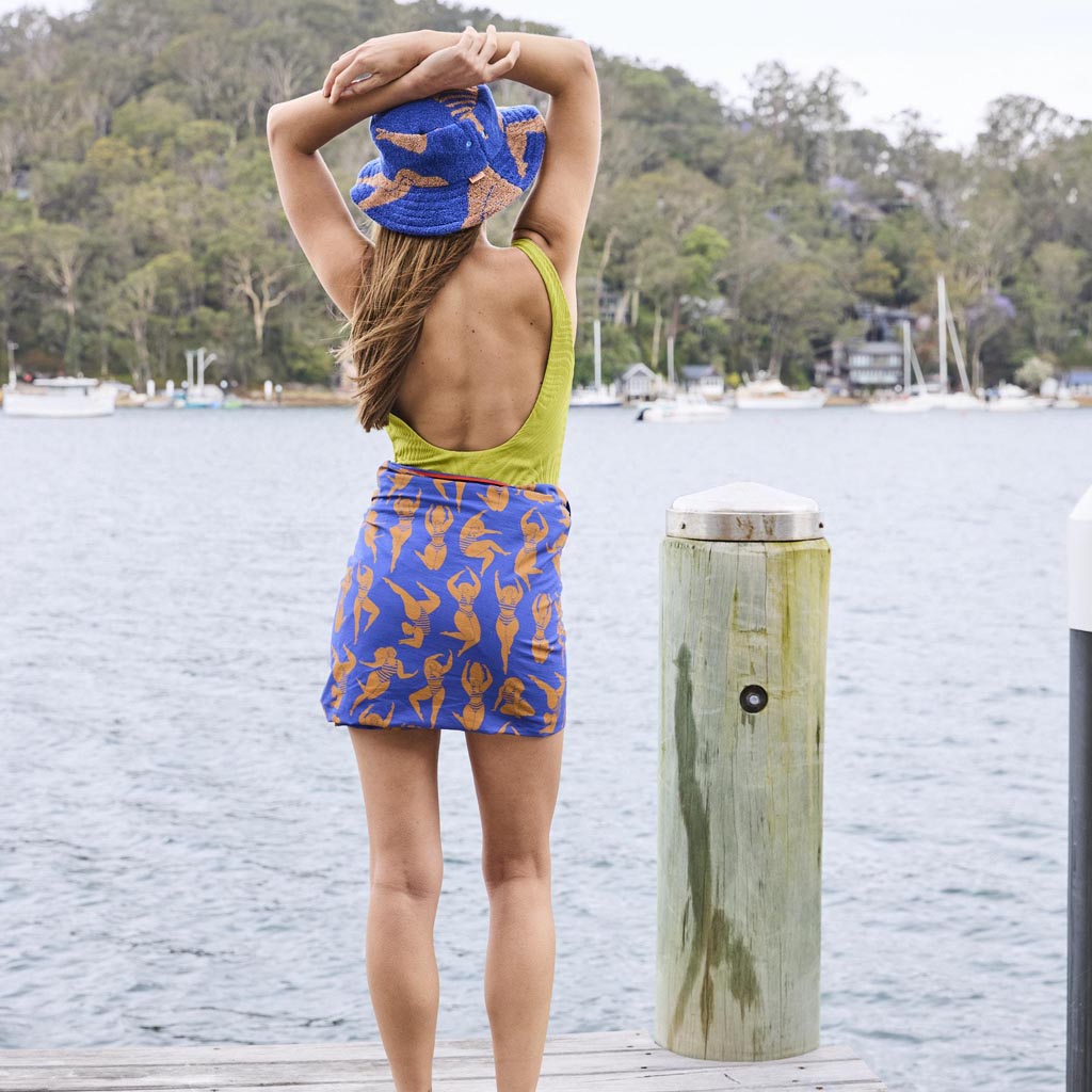 The image is of a colorful sarong made from organic cotton. The sarong features a vibrant design with a mix of warm colors, including shades of orange, pink, and yellow, reminiscent of a sun-kissed theme. The fabric appears lightweight and soft, suitable for use as a beach cover-up or a stylish wrap. The sarong is displayed against a plain background, highlighting its intricate patterns and rich hues.