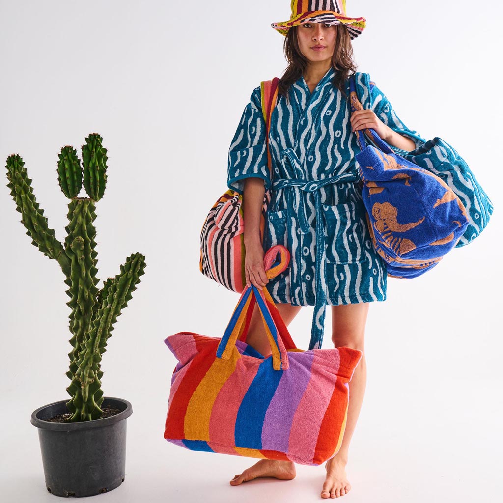 The image is of a large, colorful beach bag made from terry cloth material. The bag features a vibrant, multicolored striped pattern with shades of pink, orange, yellow, and blue, reminiscent of a sunset or a sun-kissed theme. It has sturdy handles for carrying and appears spacious, ideal for holding beach essentials. The texture of the terry cloth gives it a soft and absorbent quality, perfect for beach outings.