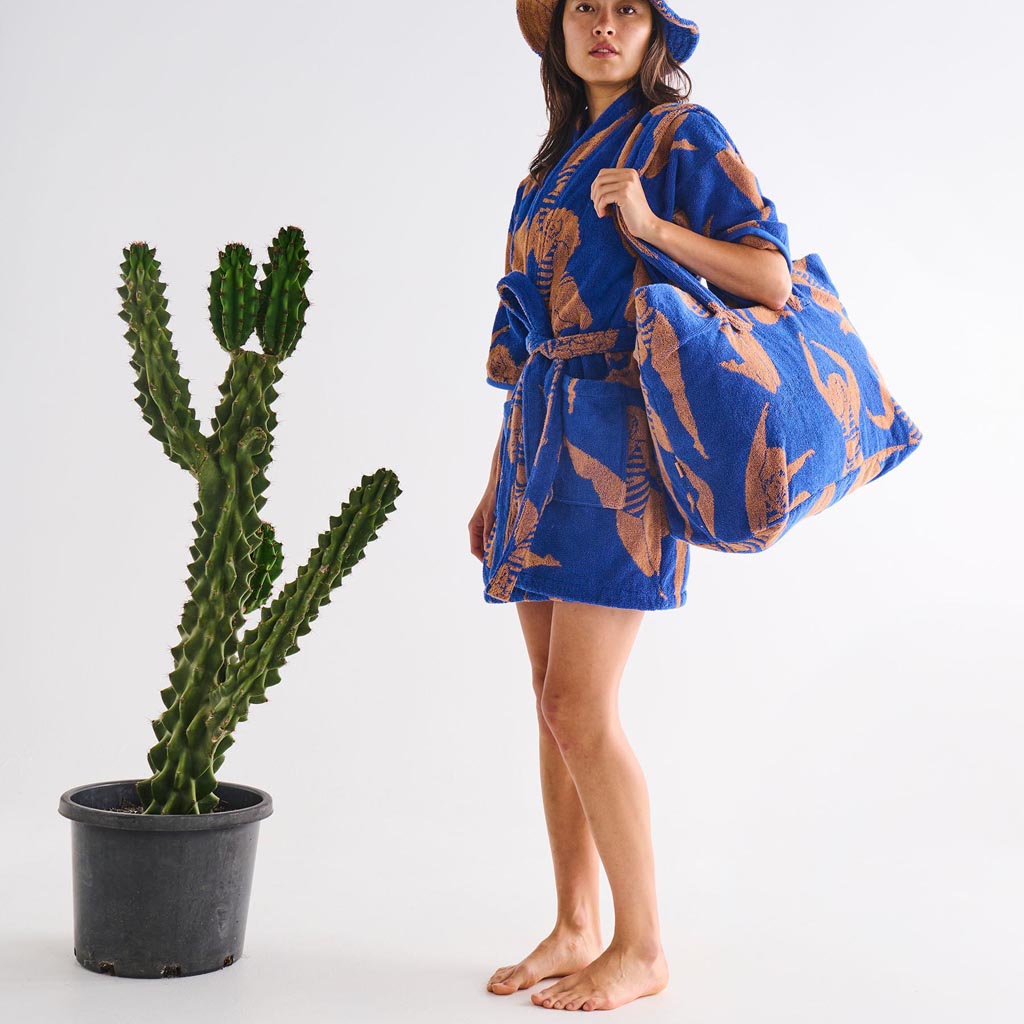 The image is of a large, colorful beach bag made from terry cloth material. The bag features a vibrant, multicolored striped pattern with shades of pink, orange, yellow, and blue, reminiscent of a sunset or a sun-kissed beach theme. The bag is oversized, suggesting it is spacious enough to carry beach essentials like towels, sunscreen, and snacks. It has sturdy handles for easy carrying, and the texture of the terry cloth gives it a soft, plush appearance. The overall design is bright and cheerful, perfect 