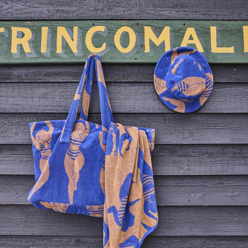 The image is of a large, colorful beach bag made from terry cloth material. The bag features a vibrant, sun-kissed design with a mix of warm colors like orange, yellow, and pink, reminiscent of a sunset or a tropical theme. The bag is oversized, suggesting it has ample space for carrying beach essentials. It has sturdy handles for easy carrying, and the texture of the terry cloth gives it a soft, plush appearance. The overall look is cheerful and perfect for a day at the beach.