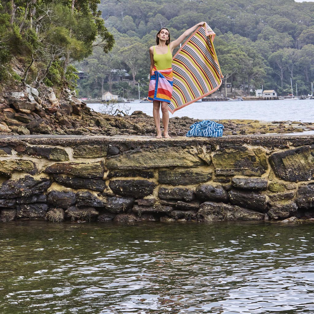 The image is of a colorful sarong made from organic cotton. The design features a vibrant and playful pattern with various abstract shapes and colors, reminiscent of a beach or surf theme. The fabric appears lightweight and is likely intended for use as a versatile beach cover-up or accessory. The background is plain, allowing the intricate details and vivid colors of the sarong to stand out prominently.