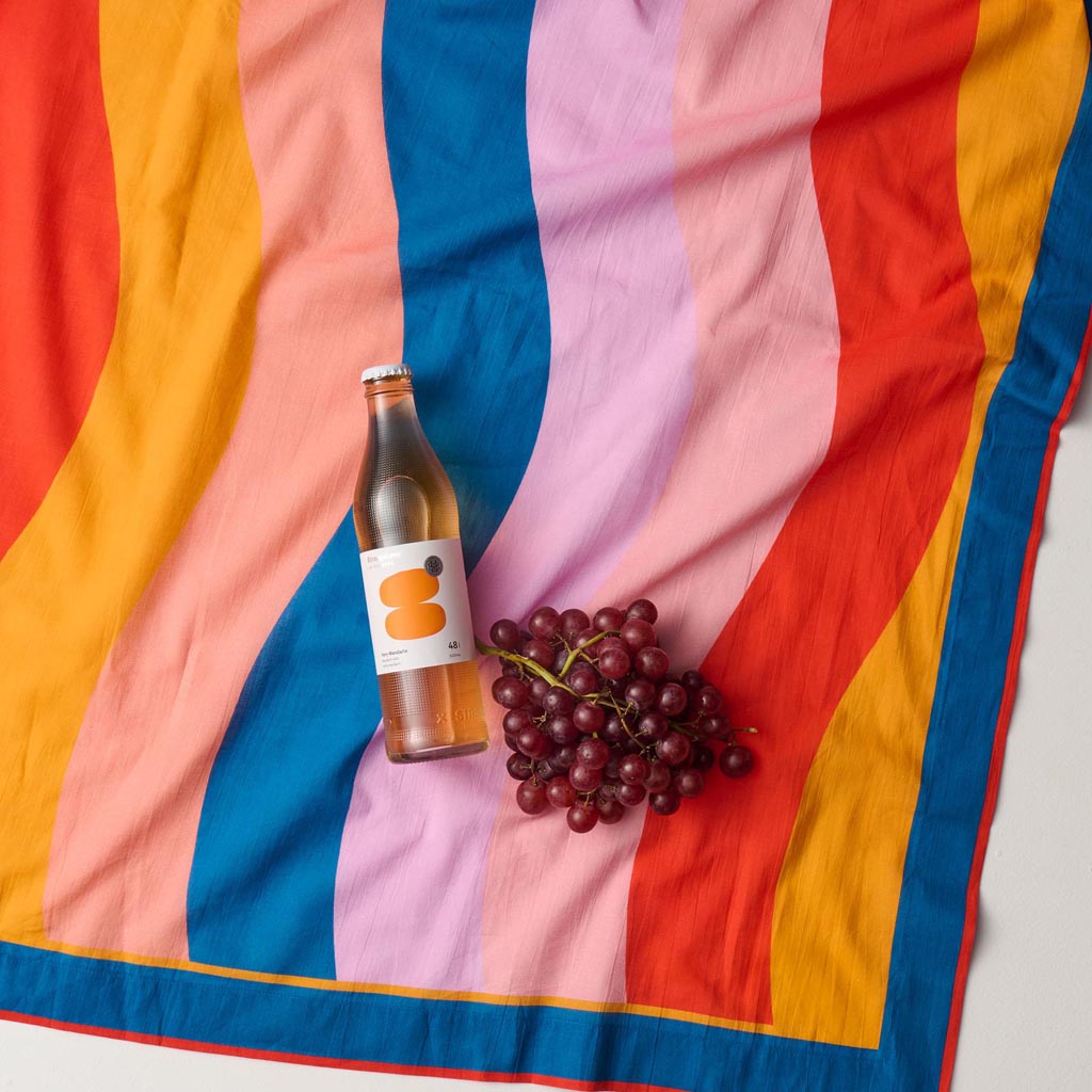 The image is of a colorful sarong made from organic cotton, designed by Kip & Co. The sarong features a vibrant and playful pattern with a mix of abstract shapes and colors, reminiscent of a beach or surf theme. The fabric appears lightweight and soft, suitable for use as a beach cover-up or a stylish accessory. The background is plain, allowing the intricate design and vivid colors of the sarong to stand out prominently.