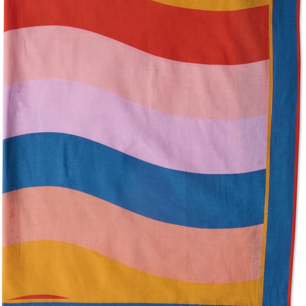 The image is of a colorful sarong made from organic cotton. The design features a vibrant and playful pattern with various shades of blue, green, and yellow, reminiscent of ocean waves or a beach scene. The fabric appears lightweight and soft, suitable for wearing as a cover-up at the beach or pool. The sarong is displayed against a plain background, highlighting its intricate design and vivid colors.