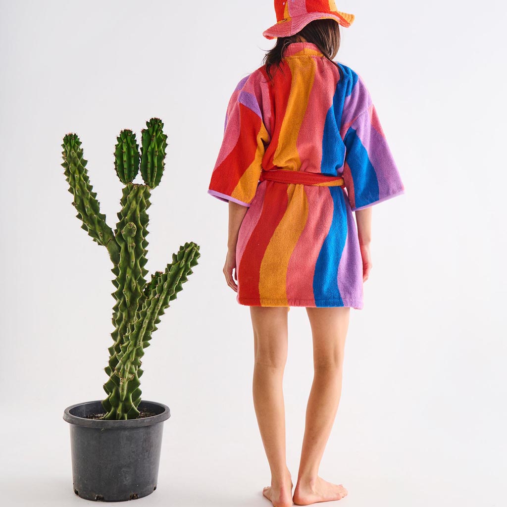 The image is of a children's robe made from terry cloth material. The robe features a colorful and playful design with a surfing theme. It includes various vibrant colors and patterns, possibly depicting surfboards, waves, or beach-related motifs. The robe has a hood, long sleeves, and a belt for tying around the waist, making it suitable for keeping warm after swimming or bathing. The overall look is fun and suitable for kids, reflecting a beach or summer vibe.