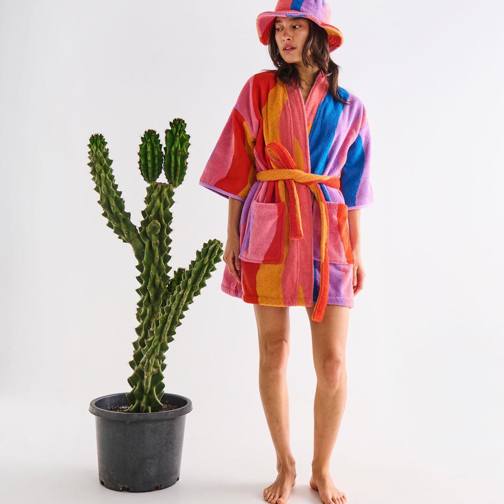 The image is of a children's robe made from terry cloth material. The robe features a colorful and playful design with a surfing theme. It includes various vibrant patterns and illustrations, such as surfboards, waves, and beach-related motifs, all in a mix of bright and cheerful colors. The robe has a hood, long sleeves, and a belt for tying around the waist, making it both functional and fun for kids.