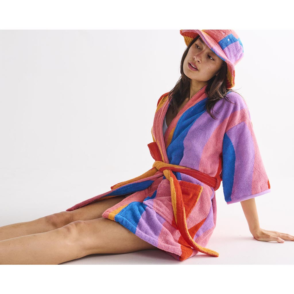 The image shows a colorful children's robe made from terry cloth material. The robe features a vibrant and playful design with various shades of blue, green, yellow, and pink, resembling a fun and lively beach or surf theme. The robe has a hood, long sleeves, and a belt for tying around the waist, making it both cozy and functional for kids. The fabric appears soft and absorbent, ideal for use after swimming or bathing. The overall look is cheerful and perfect for a beach or poolside setting.