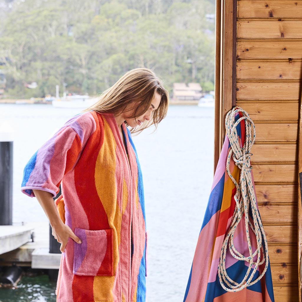 The image is of a children's robe made from terry cloth material. The robe features a colorful and playful design with a beach or surf theme, incorporating vibrant patterns and illustrations that might include surfboards, waves, or beach-related motifs. The robe has a hood, long sleeves, and a belt for tying around the waist, providing a cozy and comfortable fit for a child. The fabric appears soft and absorbent, ideal for use after swimming or bathing.