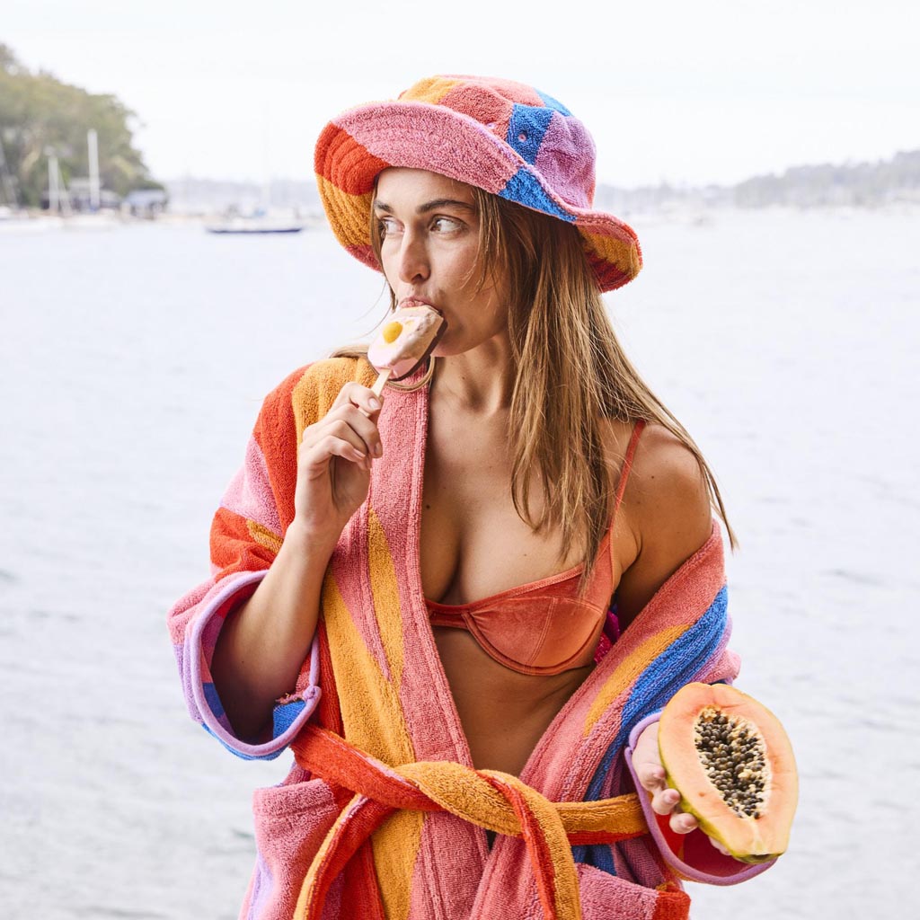 The image is of a children's robe made from terry cloth material. The robe features a colorful and playful design with a surf-themed pattern. It includes various vibrant colors and illustrations that evoke a fun, beachy vibe. The robe has a hood, long sleeves, and a belt for tying around the waist, making it both functional and cozy for kids.