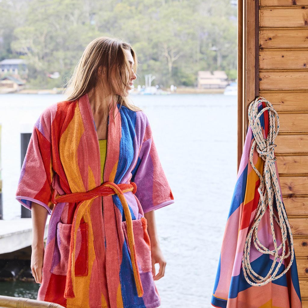 The image is of a children's robe made from terry cloth material. The robe features a colorful and playful design with various shades of blue, green, and yellow, resembling a beach or surf theme. It has a hood, long sleeves, and a belt for tying around the waist. The robe is displayed against a plain background, highlighting its vibrant pattern and soft texture.