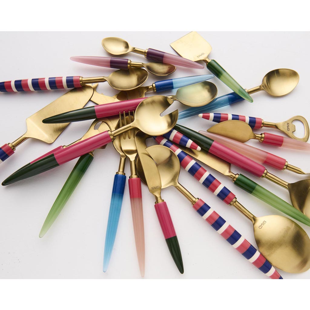 The image shows a cheese knife from the Kip & Co Tuscan Days Brasserie collection. The knife features a sleek and elegant design with a polished metal blade and a handle that appears to be made of a smooth, colorful material, possibly resin or acrylic. The handle is decorated with a vibrant, multicolored pattern that resembles a mosaic or abstract art, incorporating shades of blue, green, yellow, and red. The knife is positioned against a plain, neutral background, highlighting its artistic and functional d