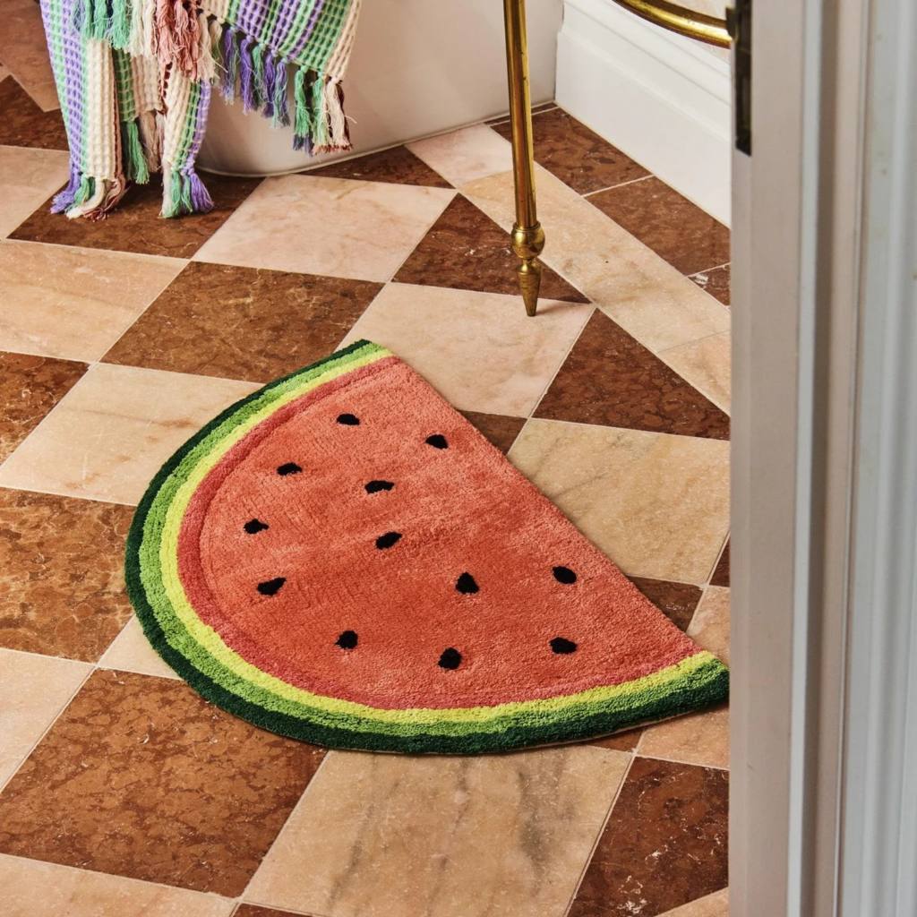 The image shows a vibrant and colorful bath mat from Kip & Co, available at Cove. The bath mat is designed in a playful watermelon pattern, featuring shades of pink and green. Made from 100% cotton, it offers a soft and comfortable feel underfoot. The mat also includes an anti-skid backing to ensure safety and prevent slipping.