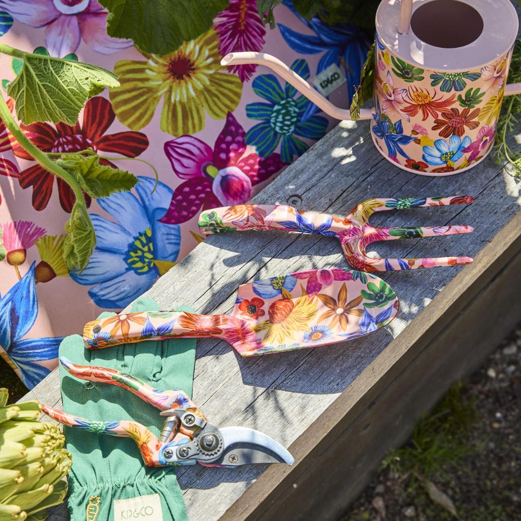 The image shows a set of garden tools and gloves with a vibrant floral design. The tools include a trowel and a hand fork, both featuring handles adorned with colorful wildflower patterns. The gloves are similarly decorated with a matching floral motif, creating a cohesive and visually appealing set. The background is likely a neutral or natural setting to highlight the bright and cheerful design of the garden tools and gloves.