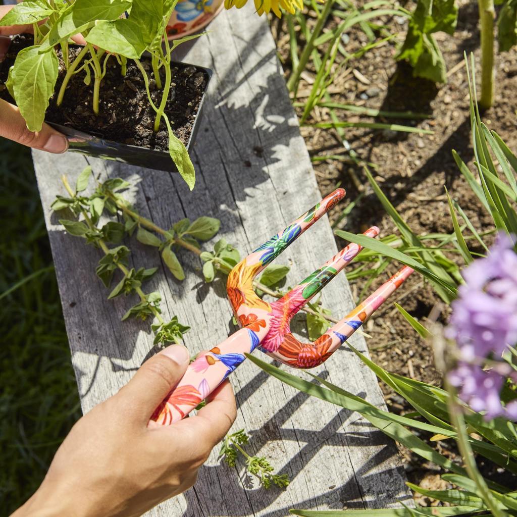 The image shows a set of garden tools and gloves with a vibrant floral design. The tools include a trowel and a hand fork, both featuring handles adorned with colorful wildflower patterns. The gloves are similarly decorated, matching the floral theme. The background is a neutral color, allowing the bright and cheerful design of the garden set to stand out. This set is likely intended for gardening enthusiasts who appreciate both functionality and aesthetic appeal in their gardening accessories.