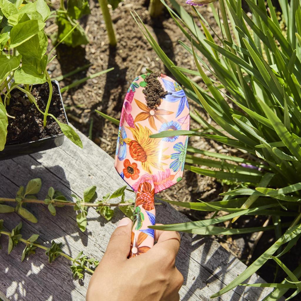 The image shows a set of garden tools and gloves with a vibrant floral design. The tools include a trowel and a hand fork, both featuring handles adorned with colorful wildflower patterns. The gloves are similarly decorated, with a matching floral motif that covers the entire surface. The background is a neutral color, allowing the bright and cheerful design of the garden set to stand out. This set is likely intended for gardening enthusiasts who appreciate both functionality and aesthetic appeal in their g