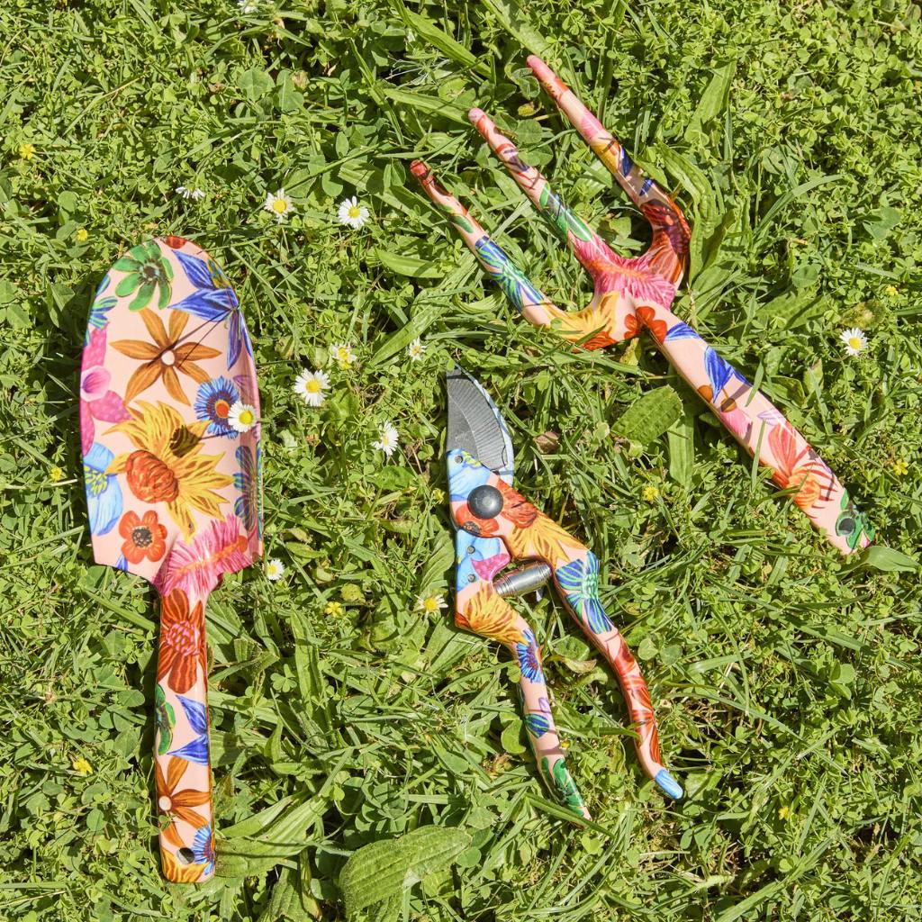 The image shows a set of garden tools and gloves with a vibrant floral design. The tools include a trowel and a hand fork, both featuring handles adorned with colorful wildflower patterns. The gloves are similarly decorated, matching the floral theme. The background is a neutral color, allowing the bright and cheerful design of the garden set to stand out. This set is likely intended for gardening enthusiasts who appreciate both functionality and aesthetic appeal in their gardening accessories.