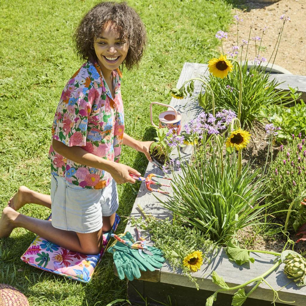 The image shows a set of garden tools and gloves with a vibrant floral design. The tools include a trowel and a hand fork, both featuring handles adorned with colorful wildflower patterns. The gloves are similarly decorated, with a matching floral motif that covers the entire surface. The background is a neutral color, allowing the bright and cheerful design of the garden set to stand out. This set is likely intended for gardening enthusiasts who appreciate both functionality and aesthetic appeal in their t
