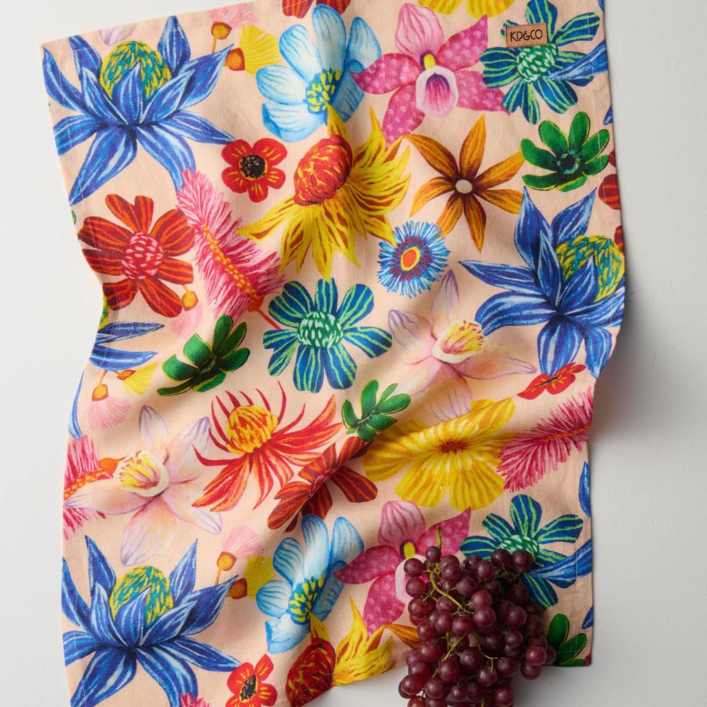 The image is of a colorful tea towel featuring a vibrant floral design. The pattern includes a variety of wildflowers in full bloom, showcasing an array of colors such as pink, yellow, blue, and green. The background of the towel is a light, neutral color that allows the bright flowers to stand out. The overall look is cheerful and lively, making it a decorative and functional kitchen accessory.