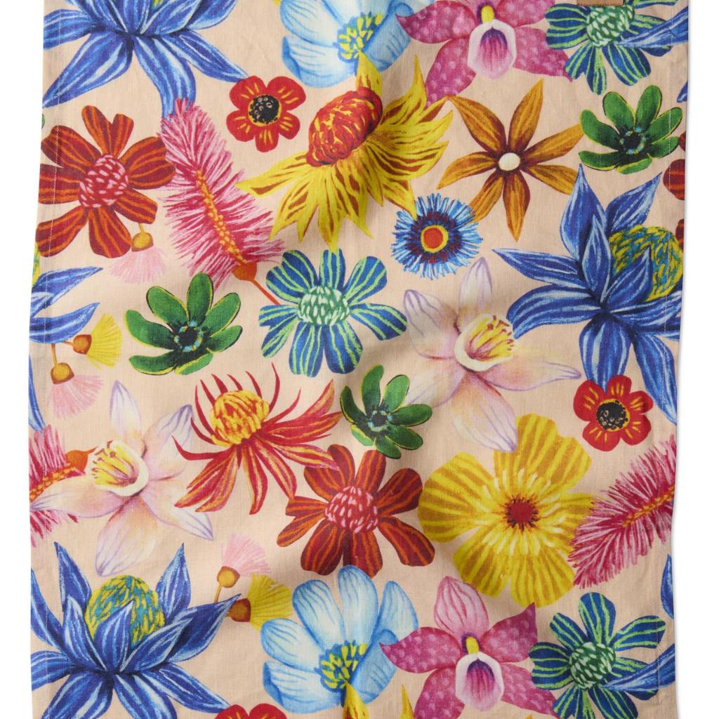 The image is of a colorful tea towel featuring a vibrant floral design. The pattern includes a variety of wildflowers in full bloom, showcasing an array of colors such as pink, yellow, blue, and green. The flowers are intricately detailed, giving the towel a lively and cheerful appearance. The background is a soft, neutral color that allows the bright flowers to stand out. The overall look is fresh and inviting, perfect for adding a touch of nature-inspired beauty to a kitchen setting.