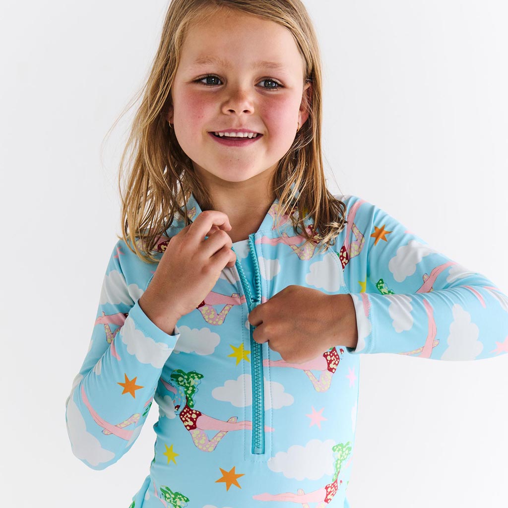 The image shows a long-sleeve bathing suit designed in collaboration between Kip & Co and The Beatles. The design is inspired by the song "Lucy In The Sky With Diamonds." The bathing suit features a vibrant and colorful pattern with psychedelic elements, likely incorporating motifs such as stars, diamonds, and abstract shapes, reflecting the whimsical and imaginative theme of the song. The background is likely a neutral color to highlight the vivid design.