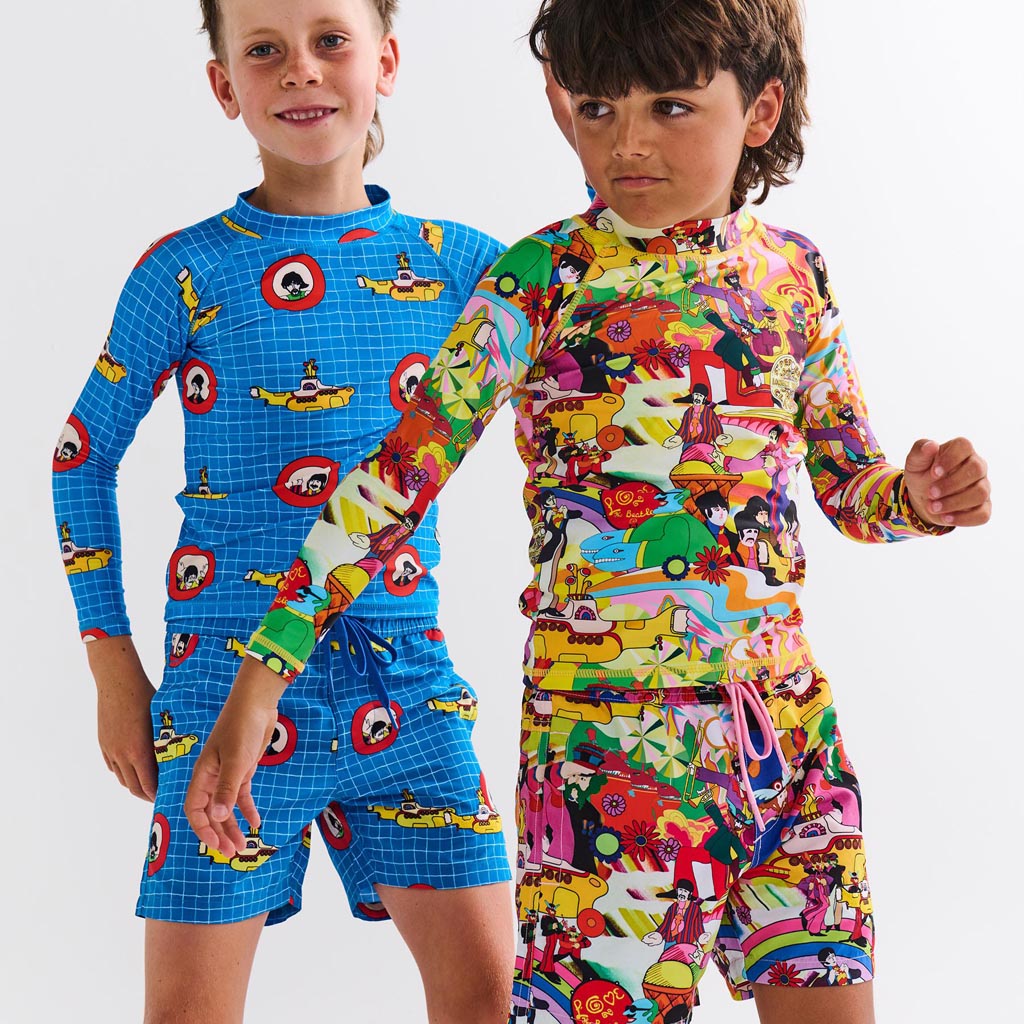 The image is likely a promotional photo for a collaboration between Kip & Co and The Beatles, featuring clothing items inspired by the iconic "Yellow Submarine" theme. The photo may show a pair of board shorts (boardies) and a rash vest, both adorned with vibrant, colorful designs reminiscent of the psychedelic art style associated with The Beatles' "Yellow Submarine" era. The clothing items might feature illustrations of the yellow submarine, characters, or other motifs from the film, set against a lively 