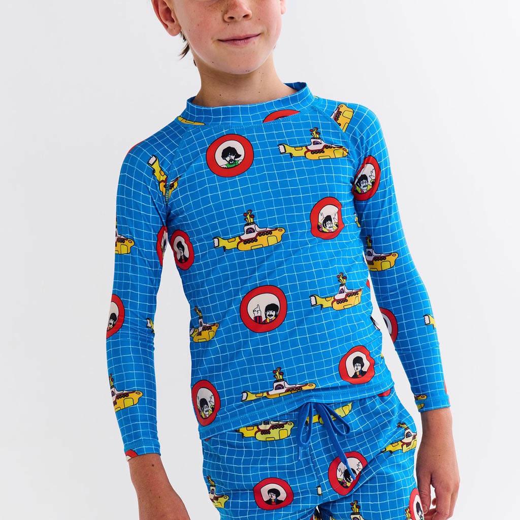 The image is likely a promotional photo for a collaboration between Kip & Co and The Beatles, featuring clothing items inspired by the iconic "Yellow Submarine" theme. The image may show a pair of board shorts (boardies) and a rash vest, both adorned with vibrant, colorful designs reminiscent of the psychedelic art style associated with The Beatles' "Yellow Submarine" era. The clothing items might feature illustrations of the yellow submarine, characters, or other motifs from the film, set against a lively 