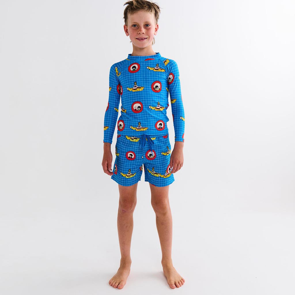 The image shows a colorful and vibrant set of children's swimwear inspired by The Beatles' "Yellow Submarine." The set includes a pair of board shorts and a rash vest. The board shorts feature a lively design with illustrations and patterns reminiscent of the psychedelic art style associated with the "Yellow Submarine" theme, incorporating bright colors like yellow, blue, and red. The rash vest complements the shorts with similar vibrant patterns and colors, creating a cohesive and playful look. The swimwea