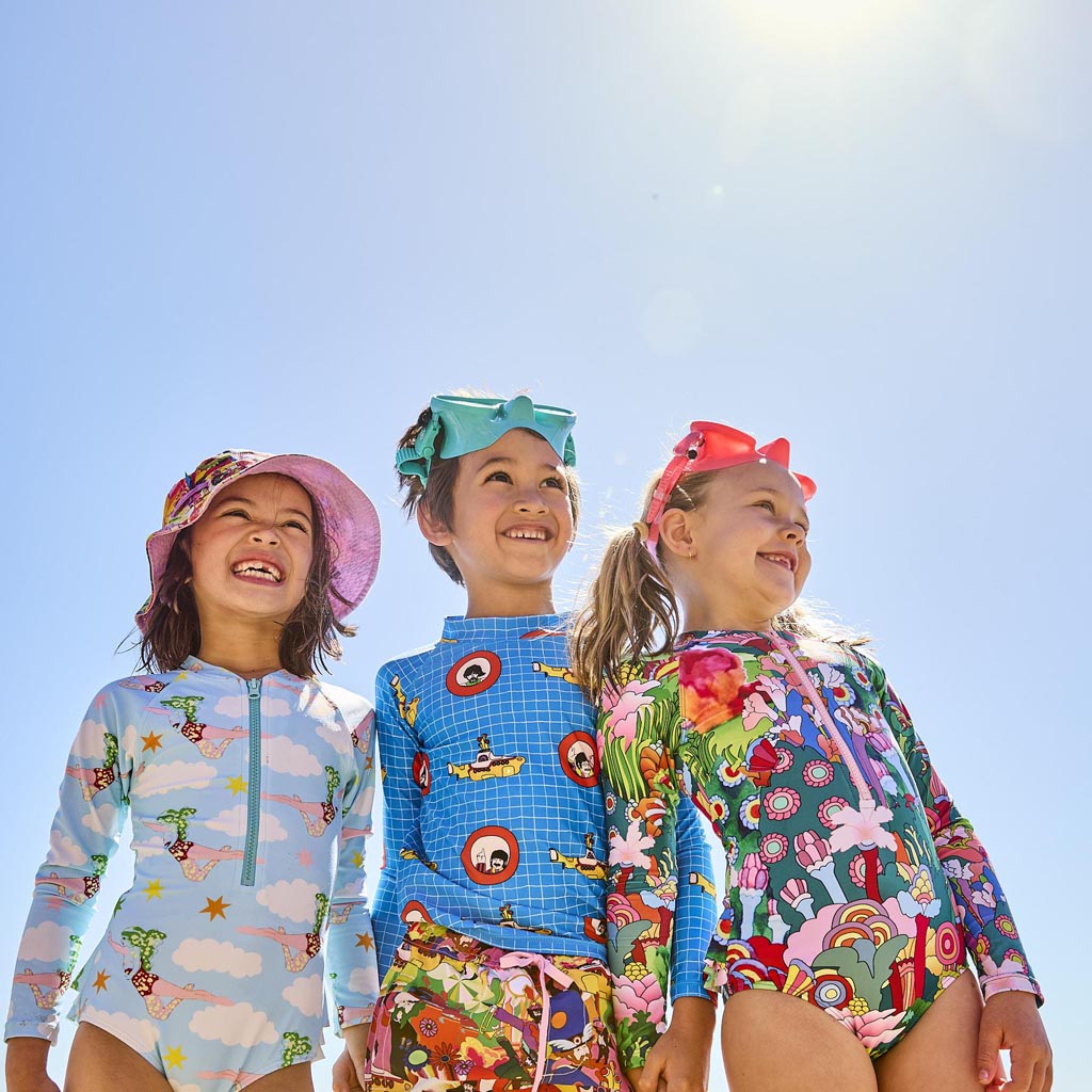The image shows a colorful and vibrant children's swimwear set inspired by The Beatles' "Yellow Submarine." The set includes a pair of board shorts and a rash vest. The board shorts feature a lively design with illustrations of the iconic yellow submarine, various sea creatures, and psychedelic patterns in bright colors like yellow, blue, and pink. The rash vest complements the shorts with similar motifs and colors, creating a cohesive and playful look. The swimwear is displayed against a plain background, 