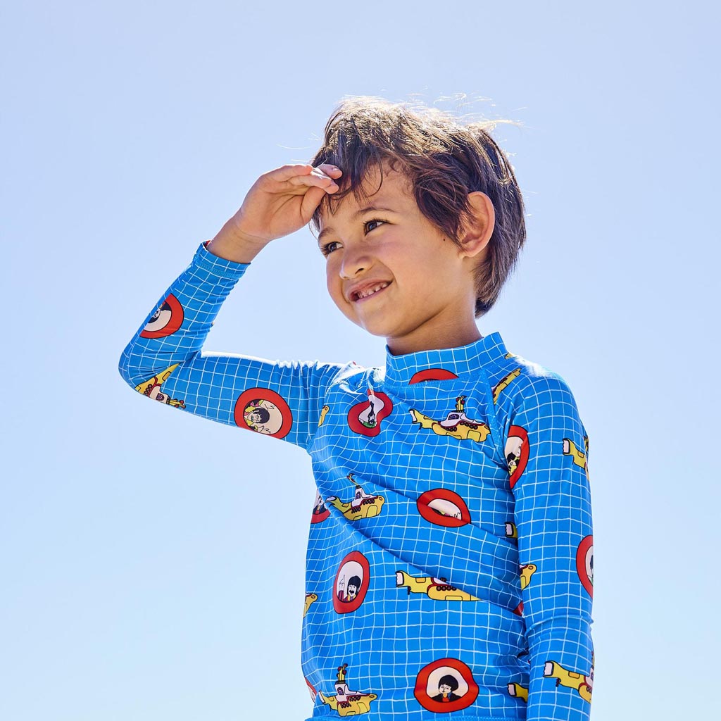 The image shows a colorful and vibrant children's swimwear set inspired by The Beatles' "Yellow Submarine." The set includes a pair of board shorts and a rash vest. The board shorts feature a lively design with illustrations of the iconic yellow submarine, various sea creatures, and psychedelic patterns in bright colors like yellow, blue, and pink. The rash vest complements the shorts with similar motifs and colors, creating a cohesive and playful look. The swimwear is laid out on a plain background, highli