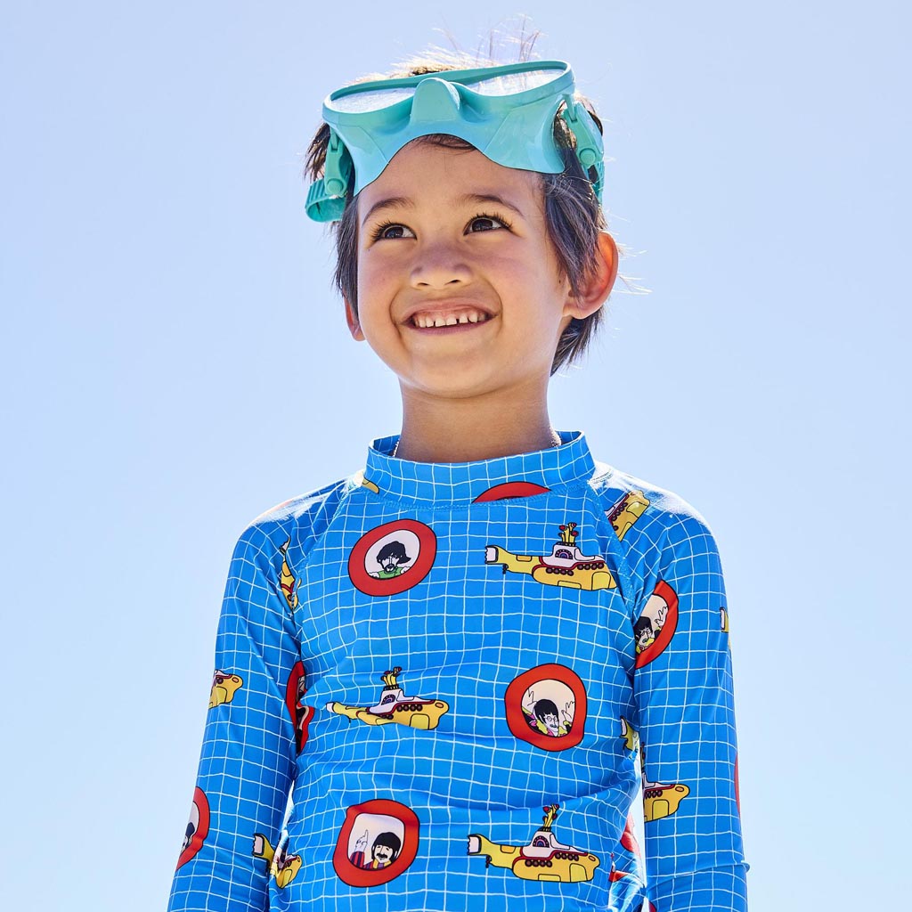 The image shows a pair of colorful board shorts designed in collaboration between Kip & Co and The Beatles, featuring a "Yellow Submarine" theme. The shorts are vibrant and eye-catching, with a pattern that includes illustrations inspired by the iconic Beatles song and animated film. The design likely incorporates elements such as submarines, sea creatures, and psychedelic patterns, reflecting the whimsical and imaginative style associated with the "Yellow Submarine" artwork.