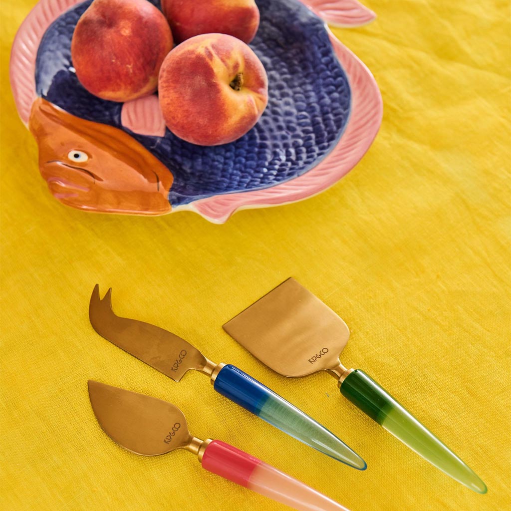 The image shows a set of three cheese knives, each with a unique and colorful handle design. The handles are adorned with vibrant, jewel-like patterns in various colors, giving them a luxurious and artistic appearance. The blades of the knives are metallic and have different shapes, each suited for cutting or serving different types of cheese. The overall presentation of the knives is elegant and eye-catching, making them not only functional but also a decorative addition to a dining setting.