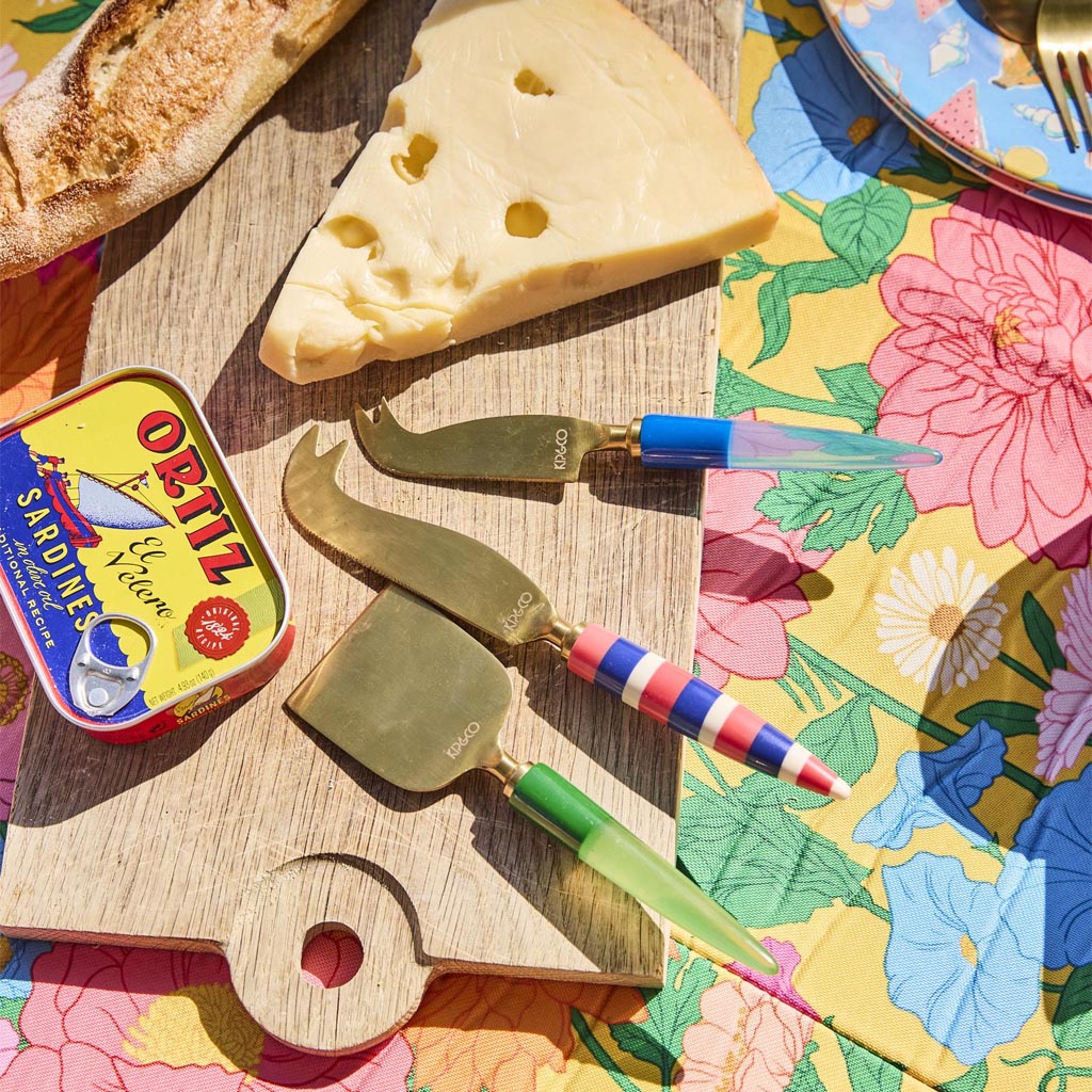 The image shows a set of three cheese knives, each with a unique and colorful handle design. The handles are adorned with vibrant, jewel-like patterns in various colors, giving them a luxurious and artistic appearance. The blades of the knives are metallic and have a sleek, polished finish. The knives are arranged side by side on a plain background, highlighting their intricate and eye-catching designs.