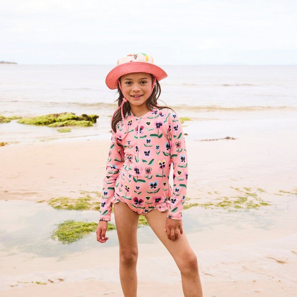 Girl on the beach in Pretty Pansy long sleeve bathers and a pink bucket hat