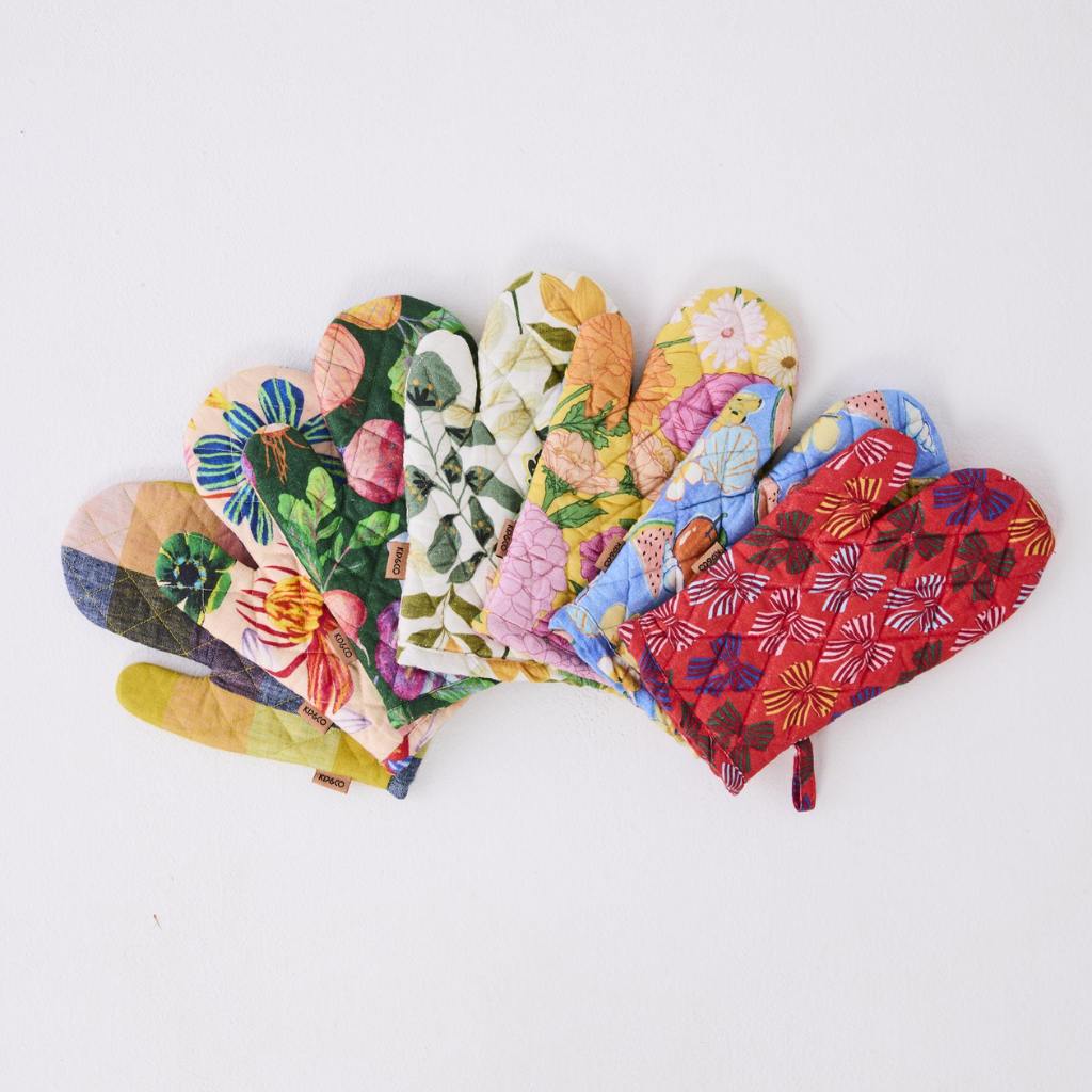 The image shows a colorful kitchen set consisting of an oven mitt and a tea towel. Both items are adorned with a vibrant vegetable soup theme, featuring illustrations of various vegetables like carrots, tomatoes, and greens, along with soup bowls and spoons. The design is playful and artistic, with a mix of bright colors that make the kitchen accessories visually appealing. The background is likely a neutral color to highlight the colorful patterns on the mitt and towel.