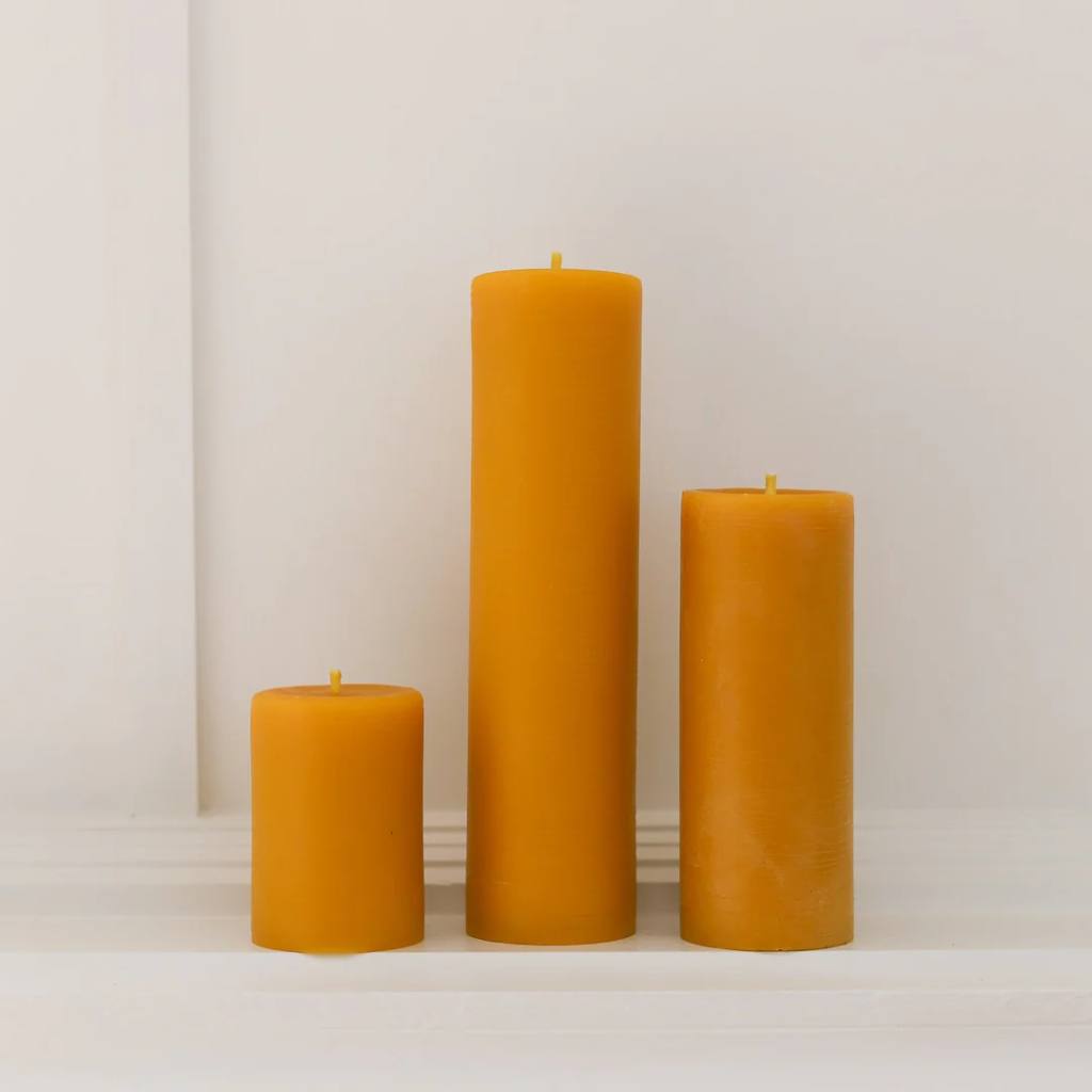 A set of large natural beeswax pillar candles from The Authentic Bee Co. displayed on a wooden surface. The candles have a smooth, creamy texture and a warm, golden hue, reflecting their natural composition. They are arranged in a visually appealing manner, showcasing their varying heights and sizes, perfect for creating a cozy and inviting atmosphere.
