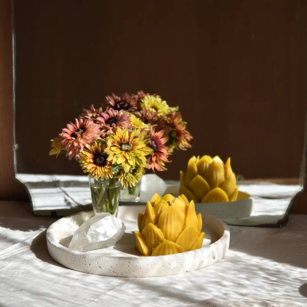 A large natural candle from The Authentic Honey Co. with a lotus design, set against a plain background. The candle appears to be made of a smooth, creamy wax, showcasing intricate details of the lotus flower.